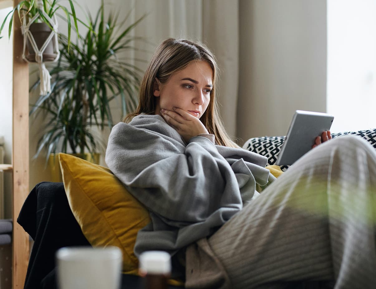 Young woman wrapped in a blanket browsing a tablet on her couch